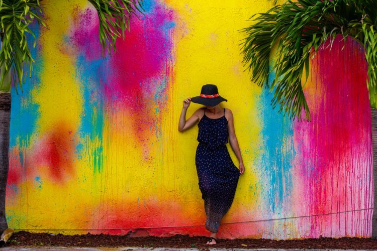 Woman in floppy hat and navy blue maxi dress standing against wall painted bright blue, pink and yellow