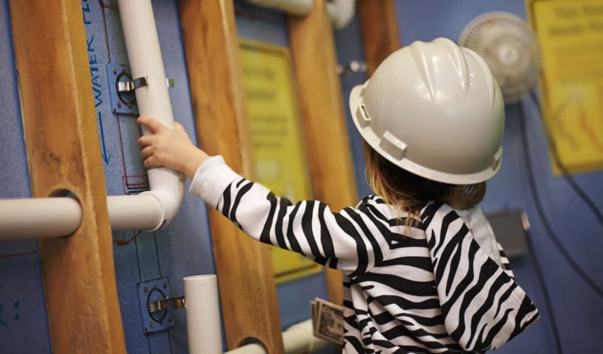 a small child wears a construction helmet and holds a pipe in a science museum