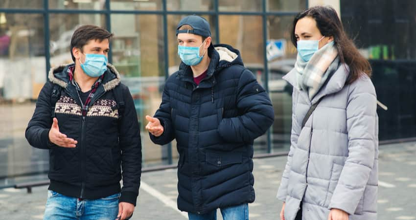 A group of three people wearing masks and winter clothes while talking on a sidewalk.