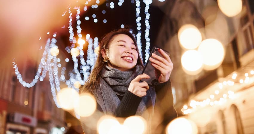 A woman smiles at her phone while surrounded by bright holiday lights