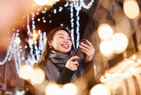 A woman smiles at her phone while surrounded by bright holiday lights