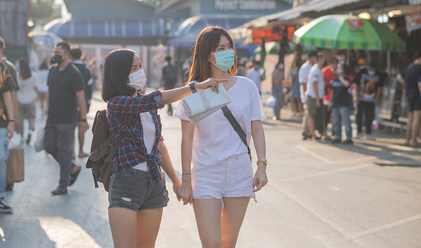 Two women hold hands and walk through a street festival, both wearing masks