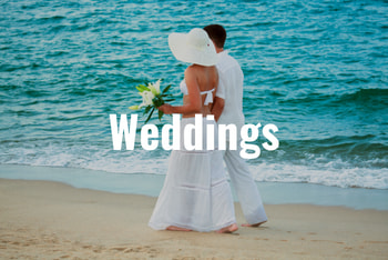 a couple in white clothes, the woman wearing a white hat and holding flowers, walks along the beach after their wedding